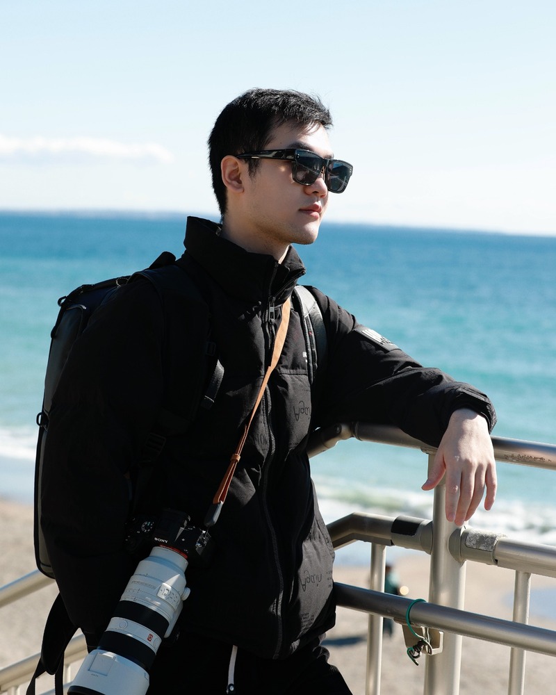 Hardy leaning on a railing by the sea with a camera and backpack